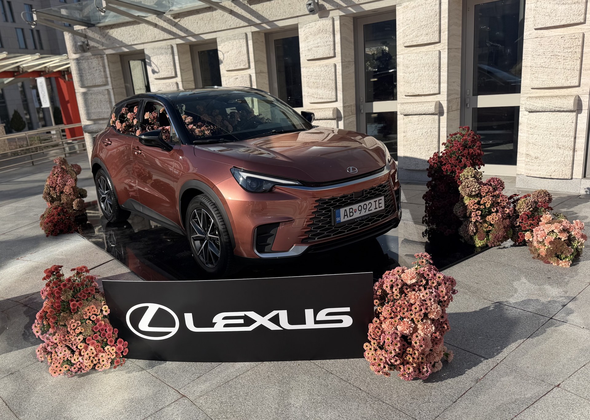 Medeno-bronzové SUV Lexus ozdobené kyticami ružových a bordových kvetov pred svetlou kamennou fasádou, v popredí čierny panel s logom Lexus
