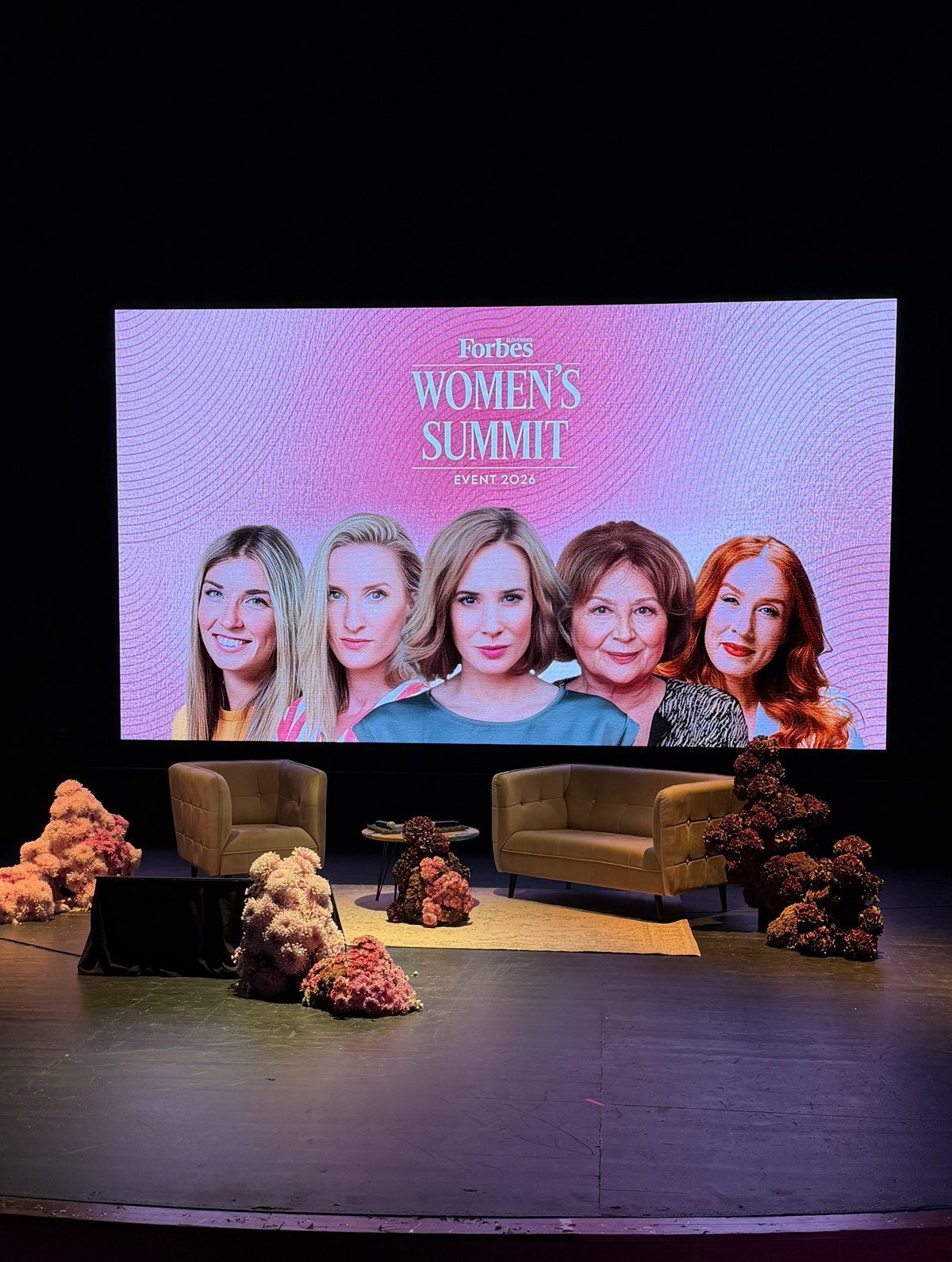 Scéna Forbes Women's Summit: kreslo, pohovka a okrúhly stolík na svetlom koberci, okolo kvetinové aranžmány na zemi a za nimi veľká LED stena s portrétmi rečníčok